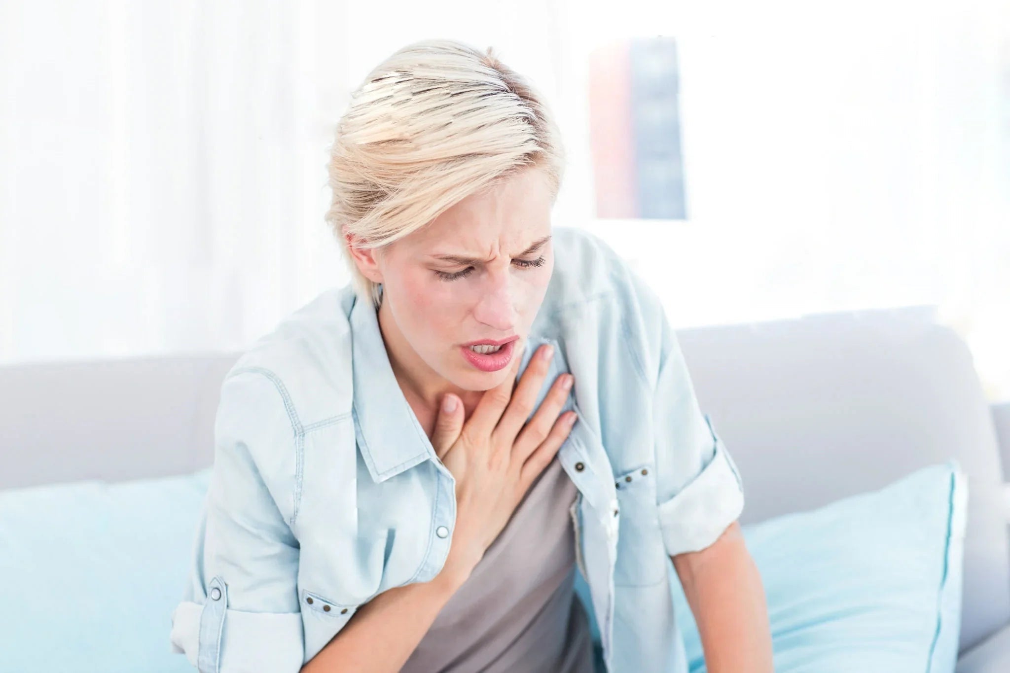 Woman with short blonde hair clutching chest, showing signs of discomfort, sitting indoors