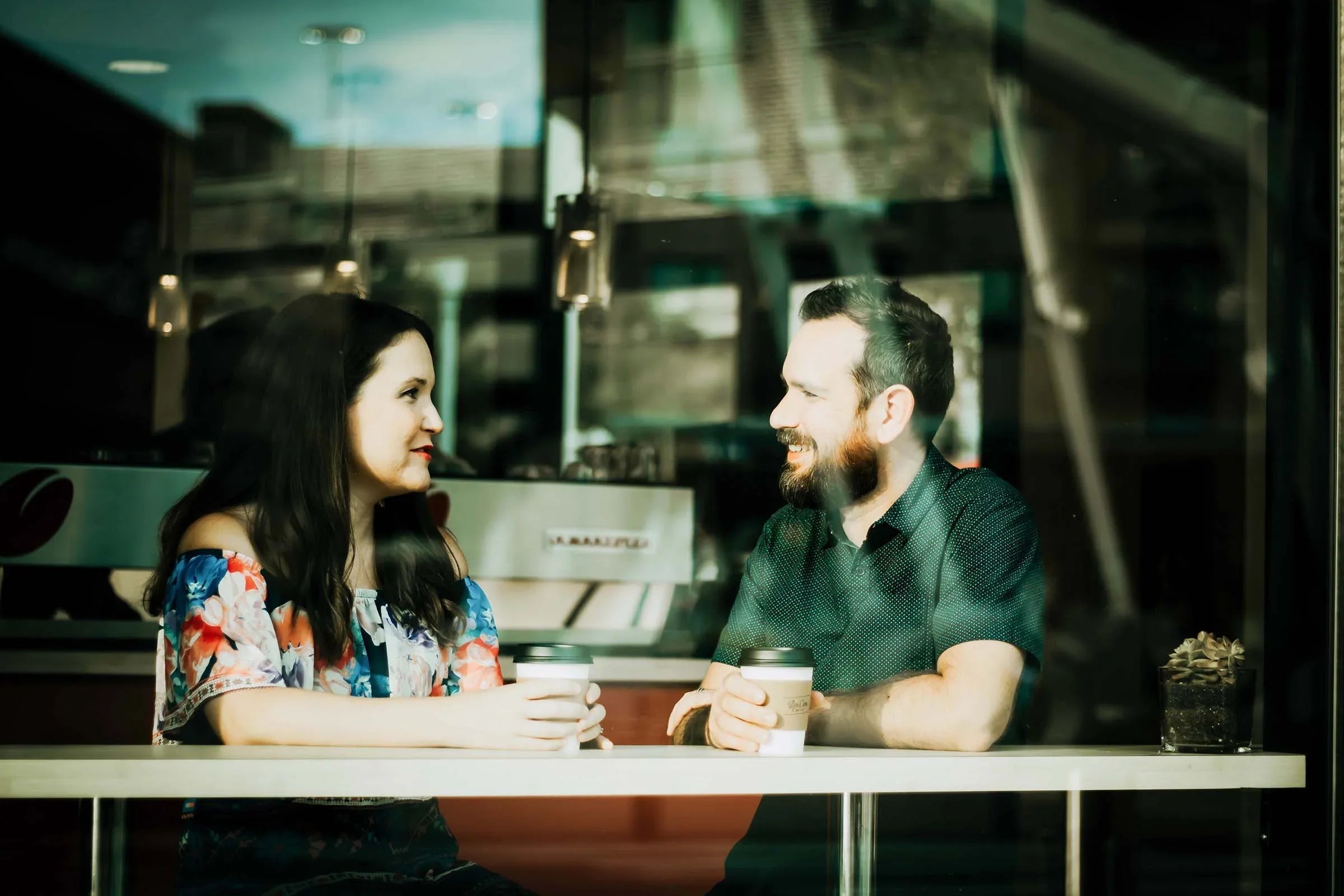 Man and woman in cotton clothing smiling and talking at a café with coffee cups