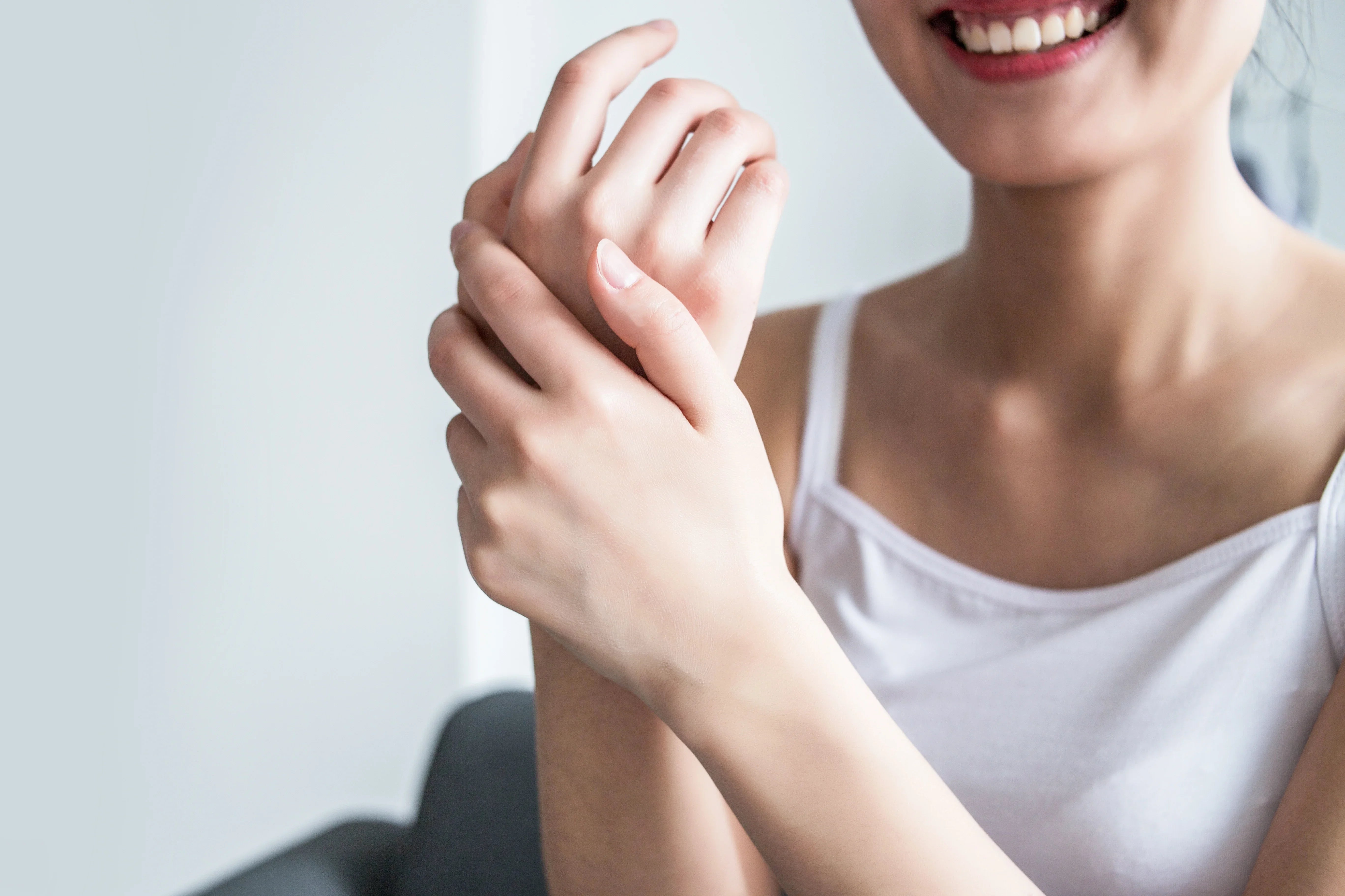 Smiling woman in white tank top applies cream to hands, promoting hypoallergenic skincare.