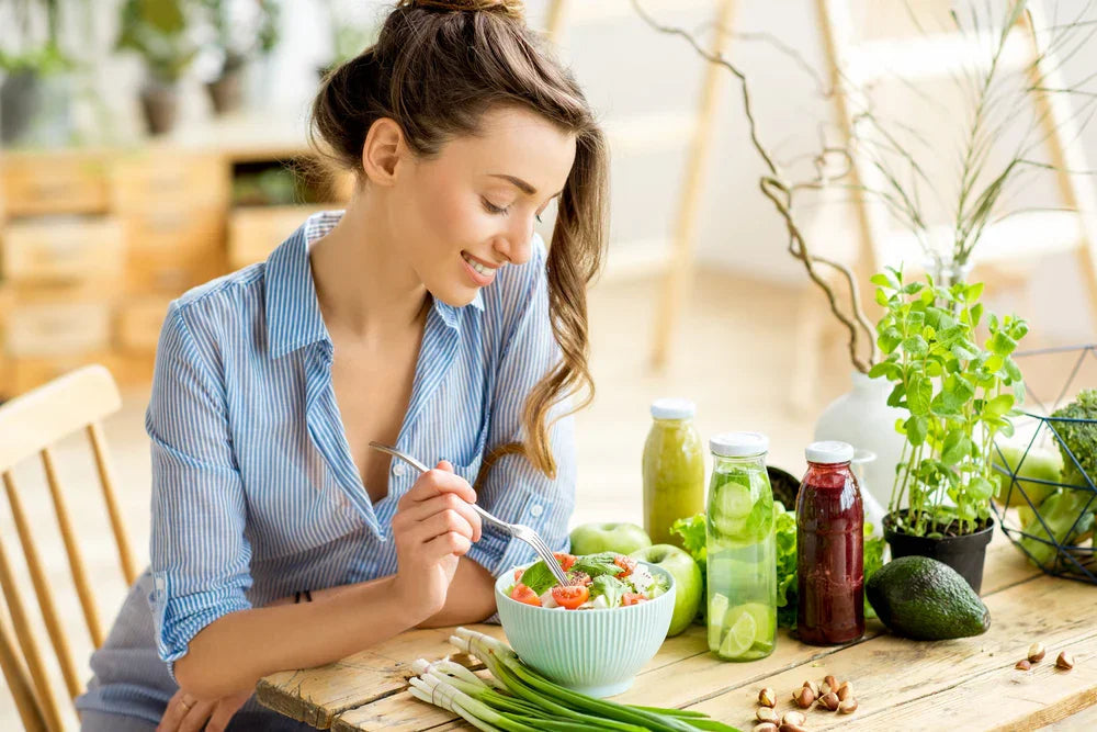 Woman in blue striped shirt eating a fresh salad, surrounded by green juices, avocado, and herbs, highlighting allergy-free, healthy lifestyle.