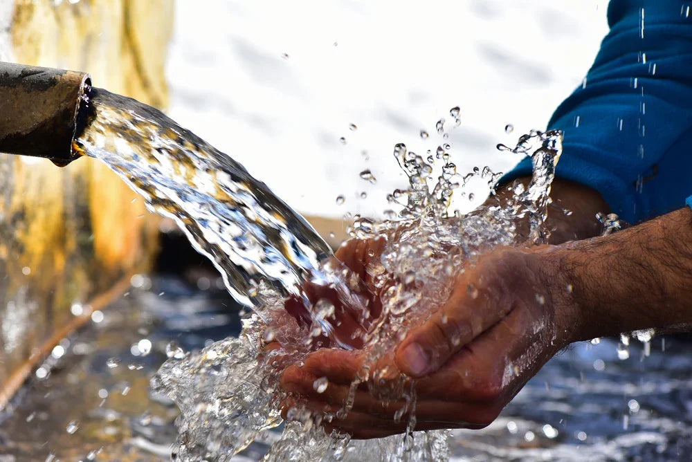 Hands in blue sleeves cupping clear flowing water from metal pipe, allergy-free apparel context.