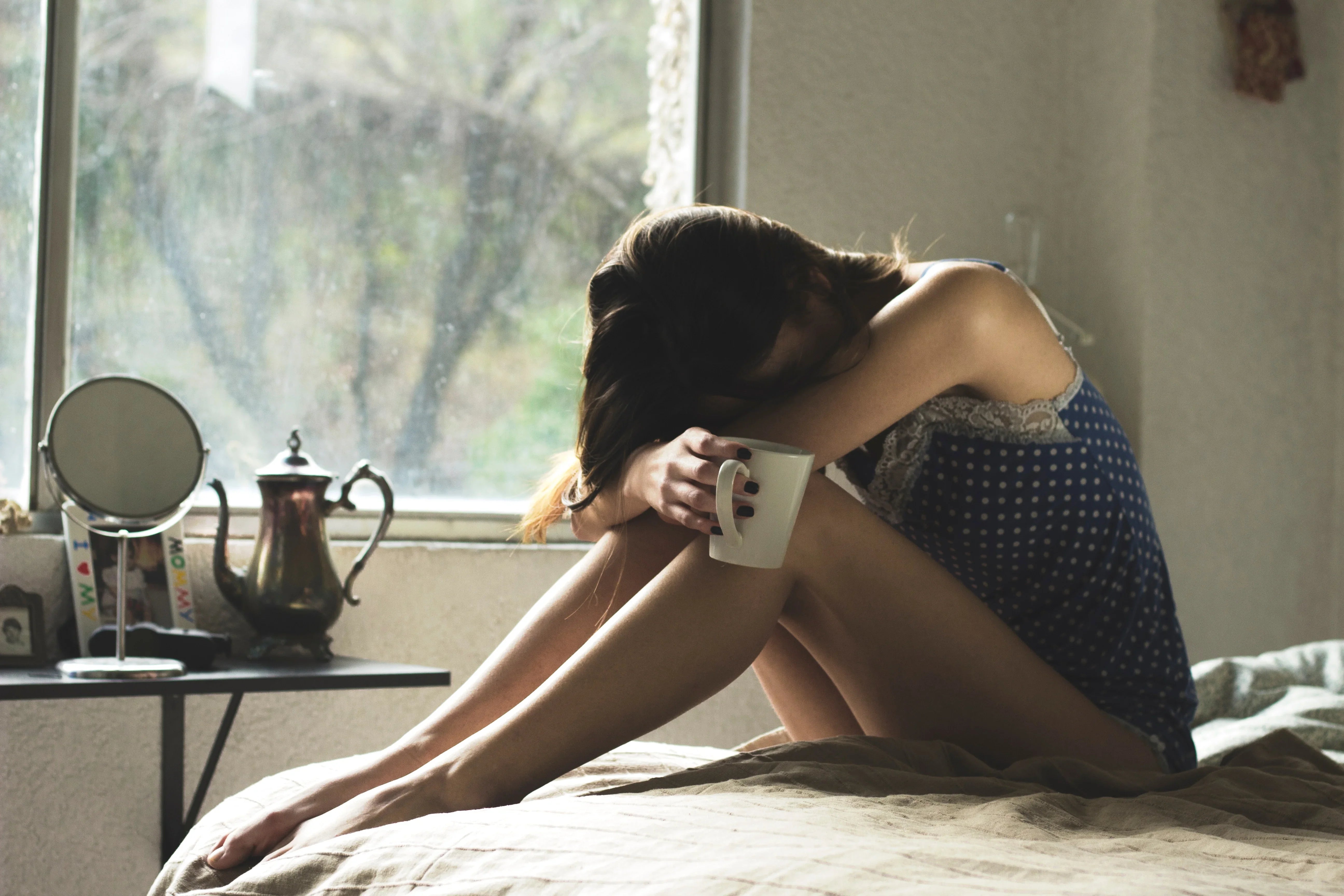 Woman in blue polka dot sleepwear sitting on bed holding coffee mug in morning light