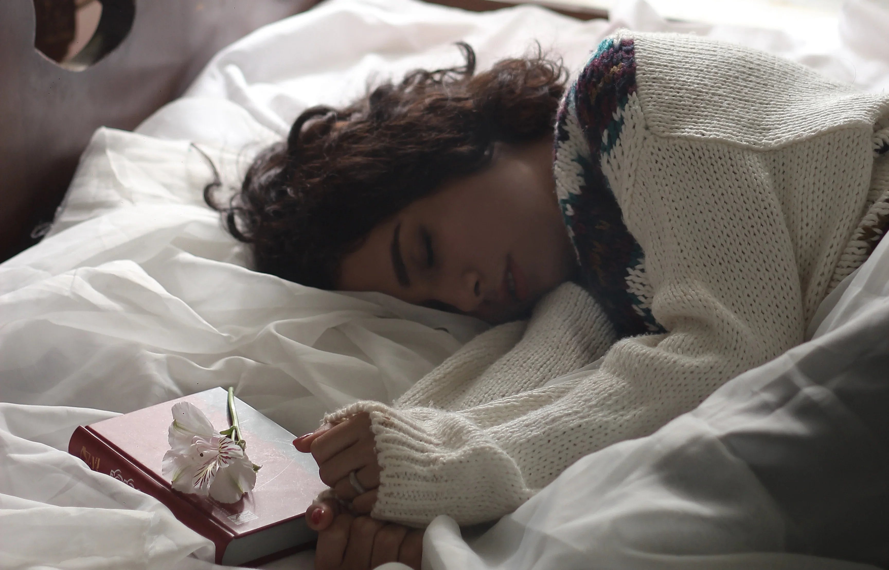Woman in hypoallergenic cotton sweater sleeps on white bedding beside a book and flower