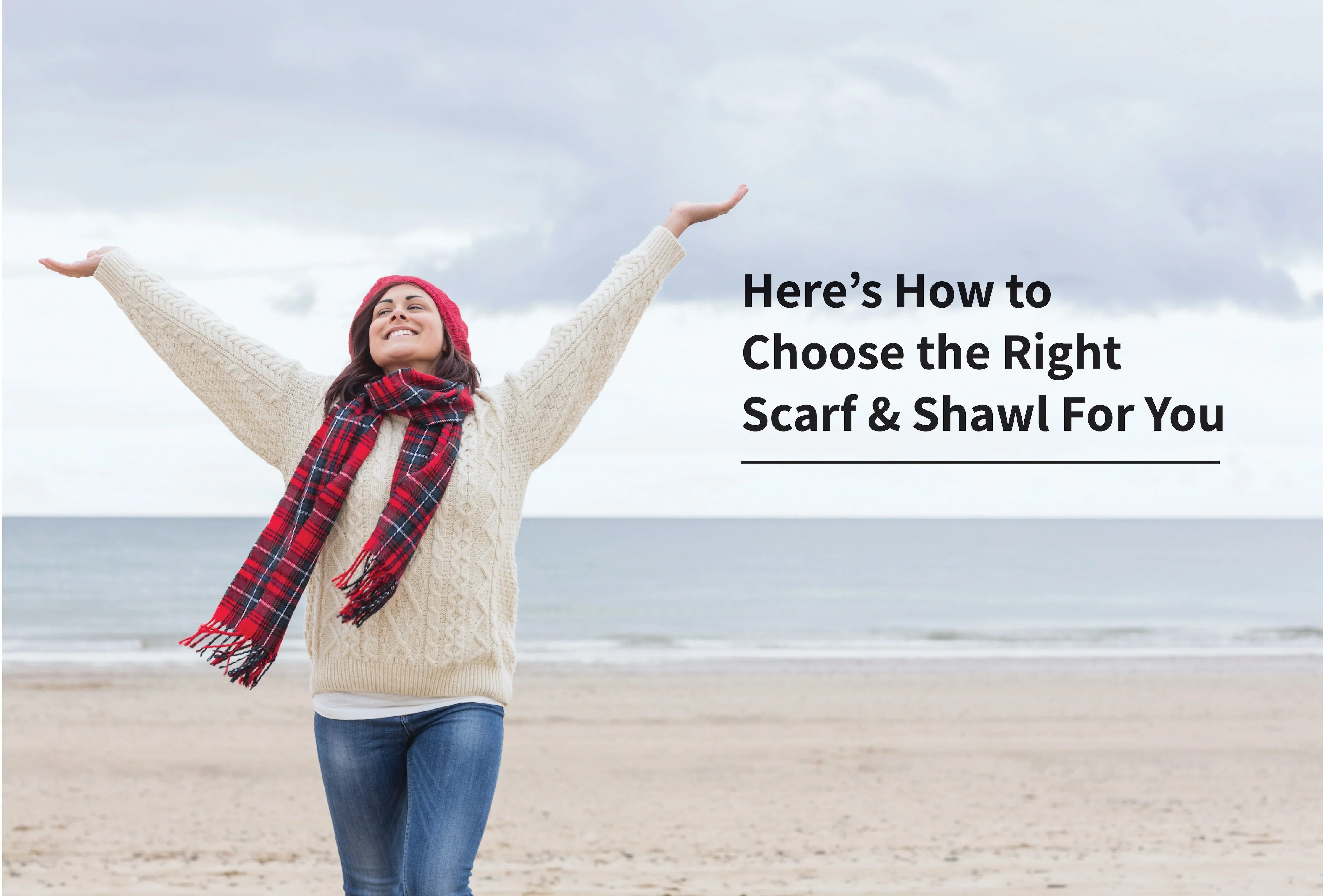 Woman in knit sweater and red plaid scarf standing on beach with arms raised, winter accessories