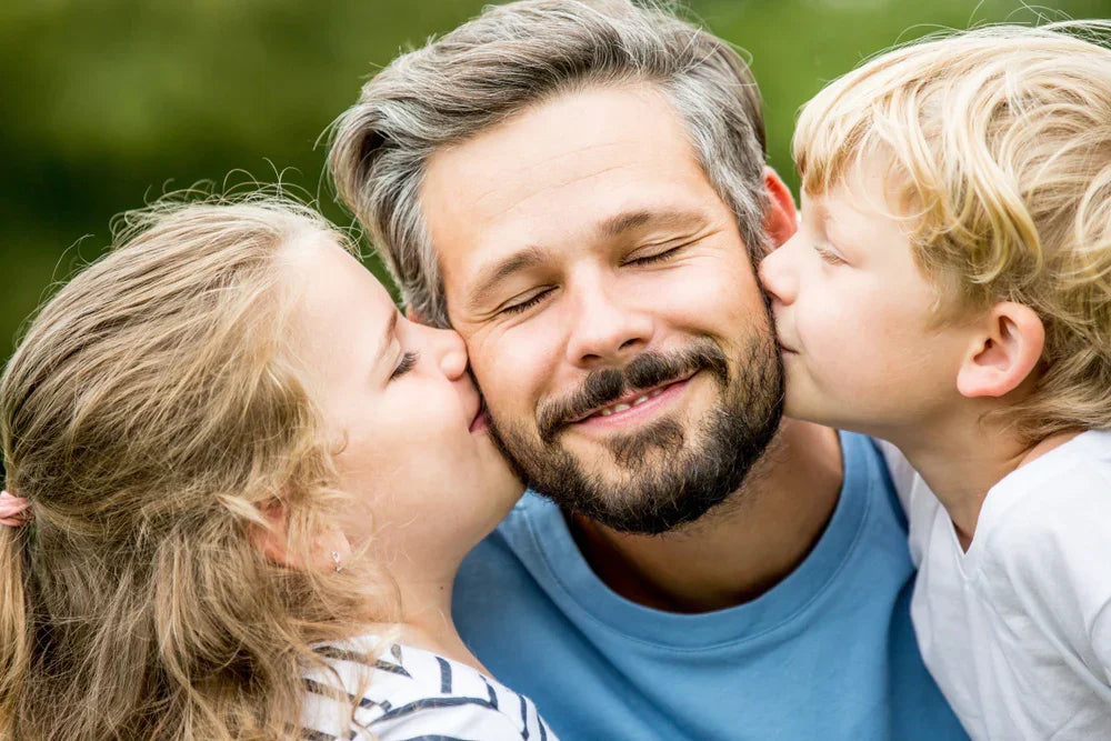 Smiling father with beard being kissed on cheeks by two kids outdoors