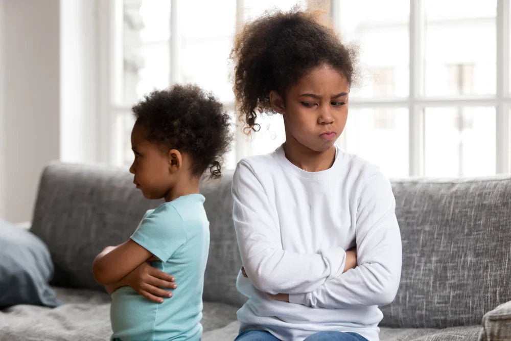 Two kids with crossed arms sitting on a couch in soft cotton shirts, looking upset