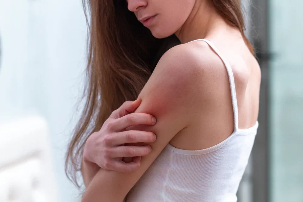 Woman scratching irritated skin on arm, wearing white tank top, sensitive skin concern