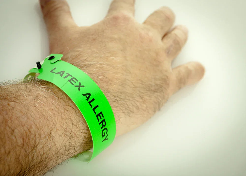 Hand wearing green wristband labeled latex allergy, allergy warning, sensitive skin alert