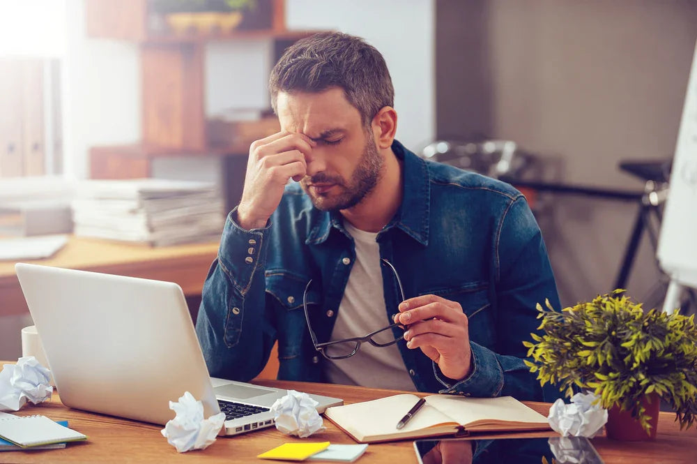 Stressed man in casual shirt at desk with laptop, notebook, and crumpled papers