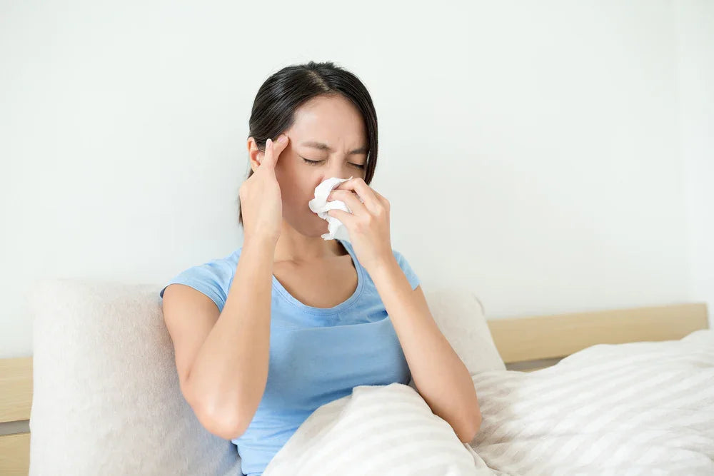 Woman in bed holding tissue to nose, showing allergy symptoms, wearing light blue cotton shirt
