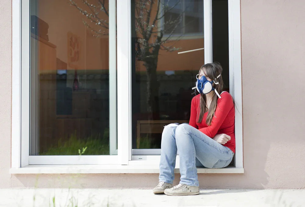 Woman in red shirt and jeans wearing an allergy mask sitting by a window, hypoallergenic apparel
