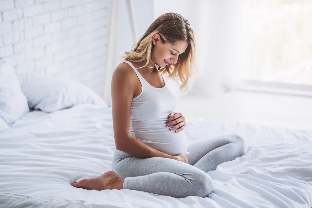 Pregnant woman wearing soft organic cotton clothing sitting on a bed in a bright bedroom