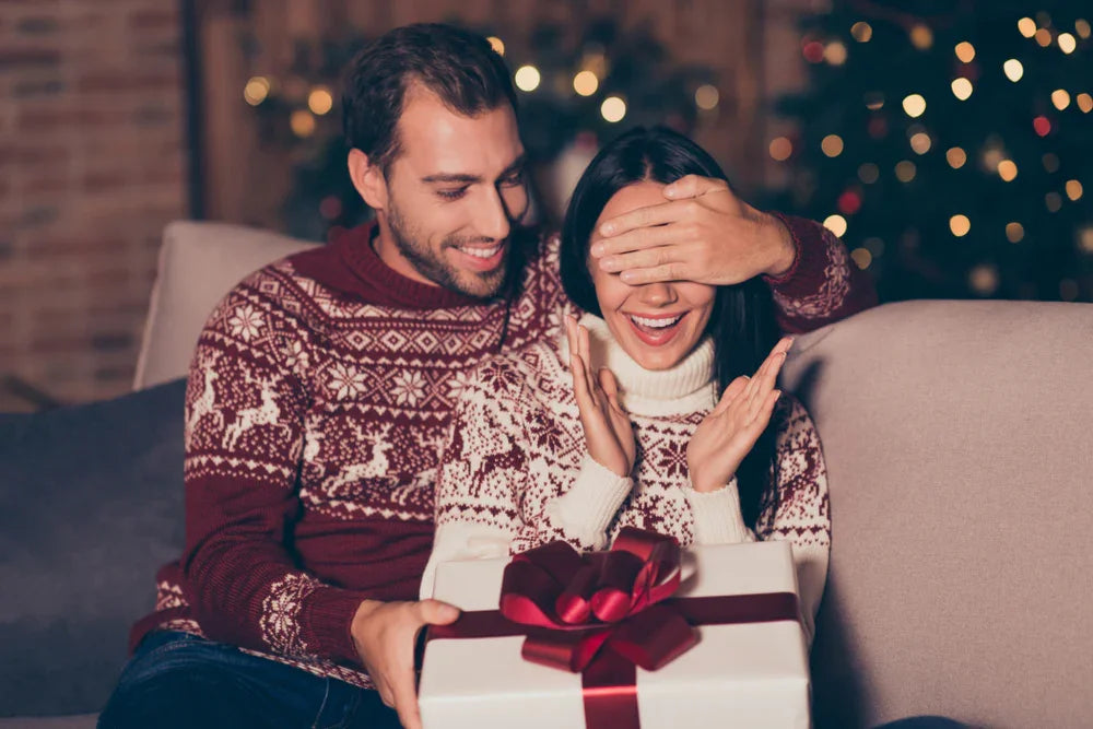 Smiling couple in matching allergy-free cotton sweaters exchanging a Christmas gift on a couch