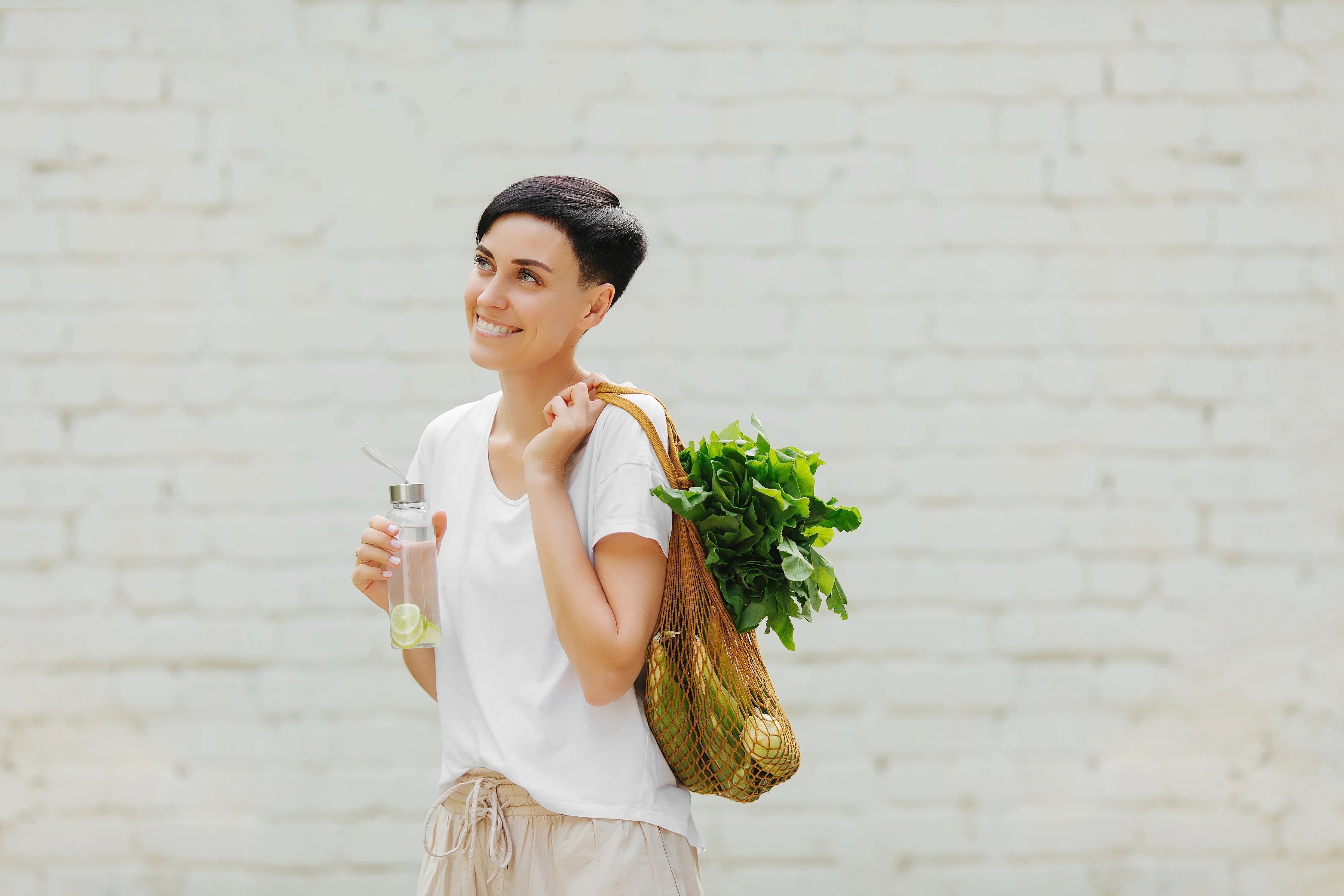 Woman in white organic cotton shirt holding reusable bag with vegetables and water bottle