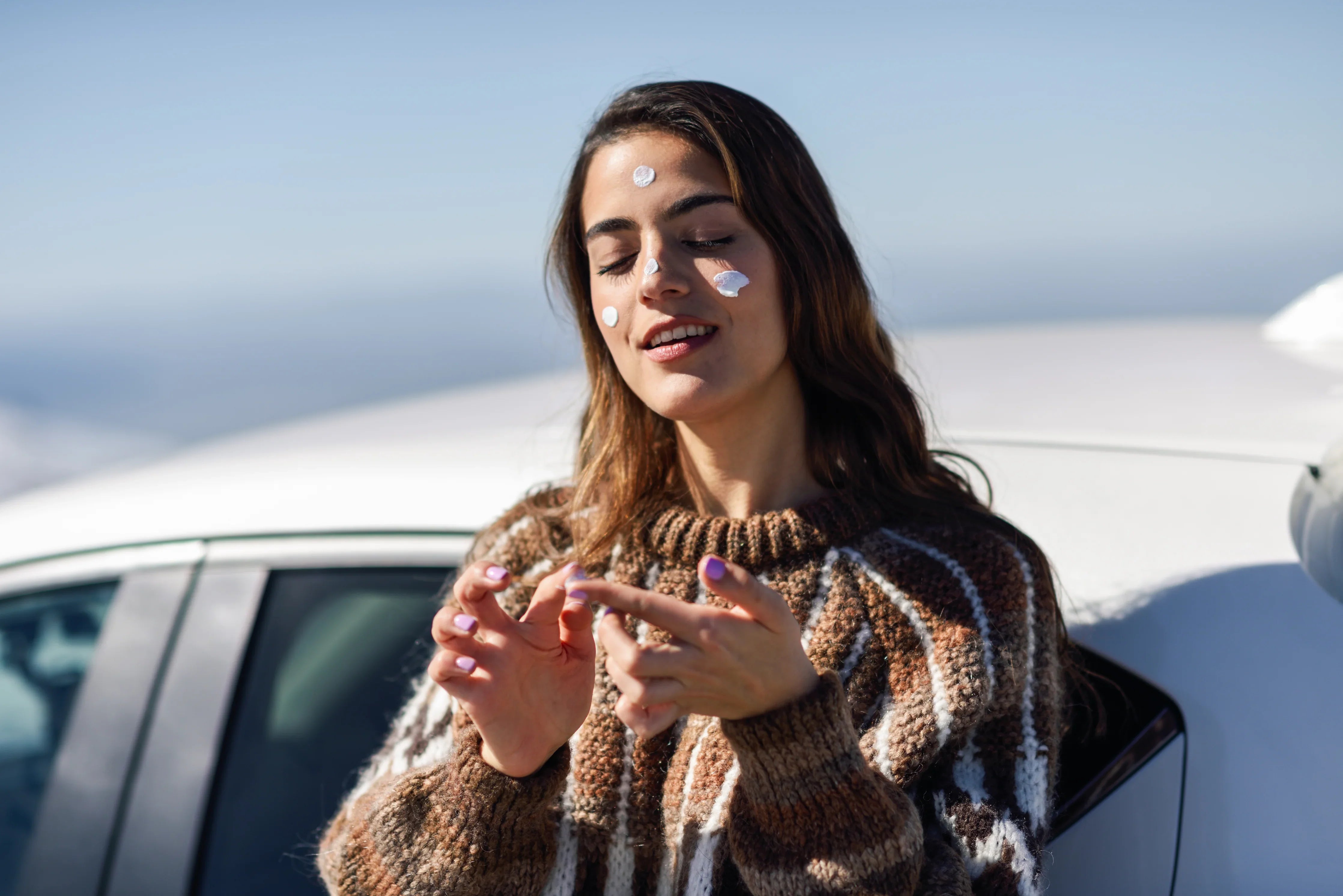 Woman in hypoallergenic knit sweater applying cream to face outdoors, beside white car