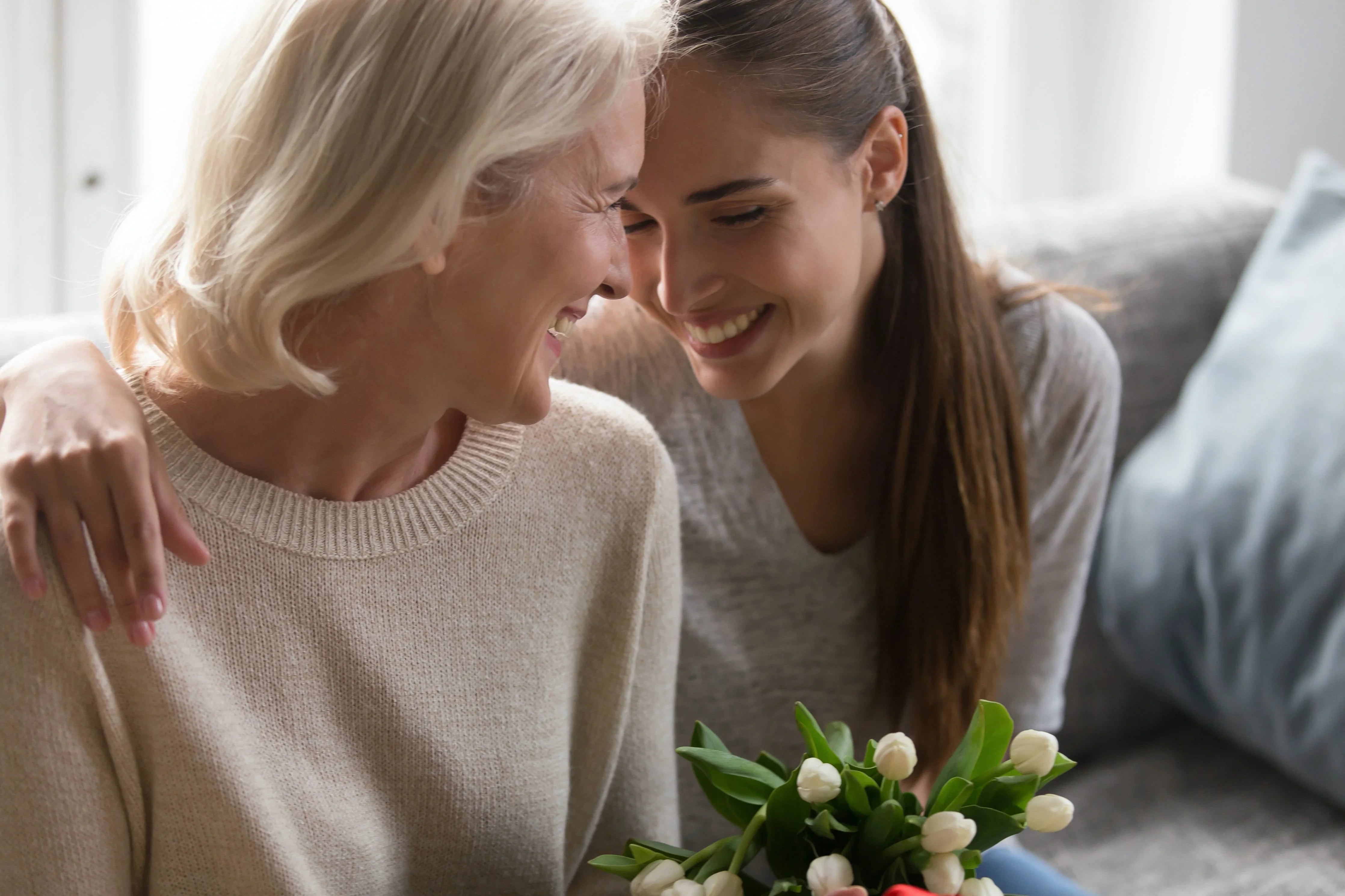 Smiling women in soft cotton sweaters embracing on sofa with white tulip bouquet