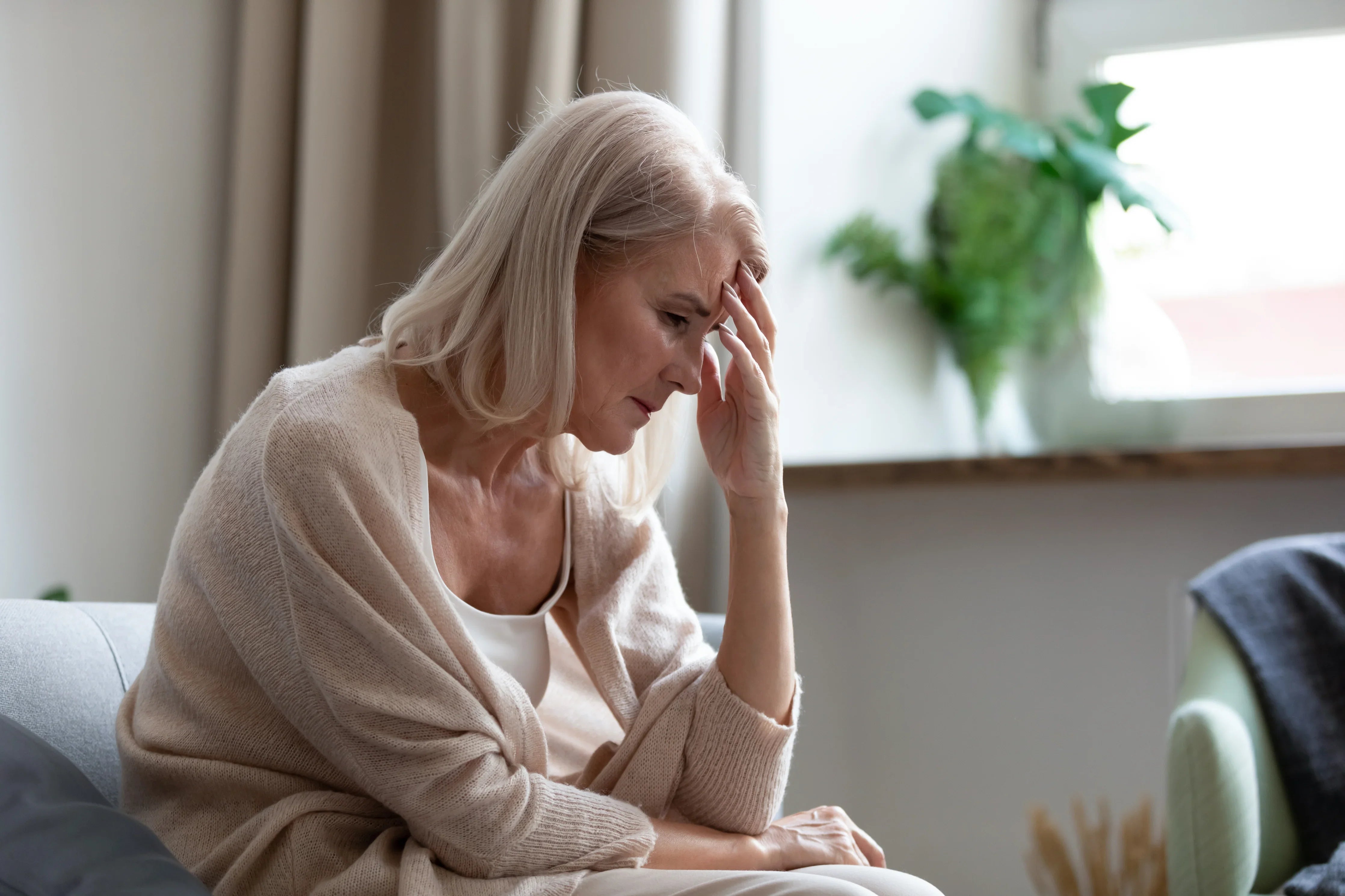 Senior woman in beige hypoallergenic sweater looking worried, sitting indoors near window