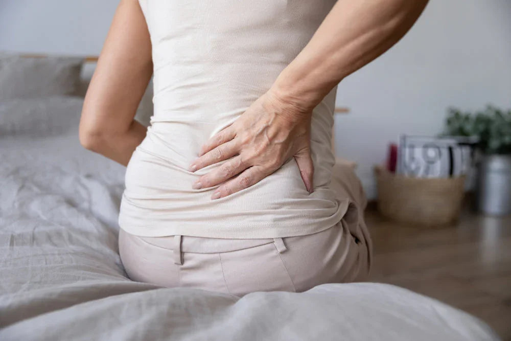 Woman wearing beige hypoallergenic cotton shirt sitting on bed, holding lower back in pain.