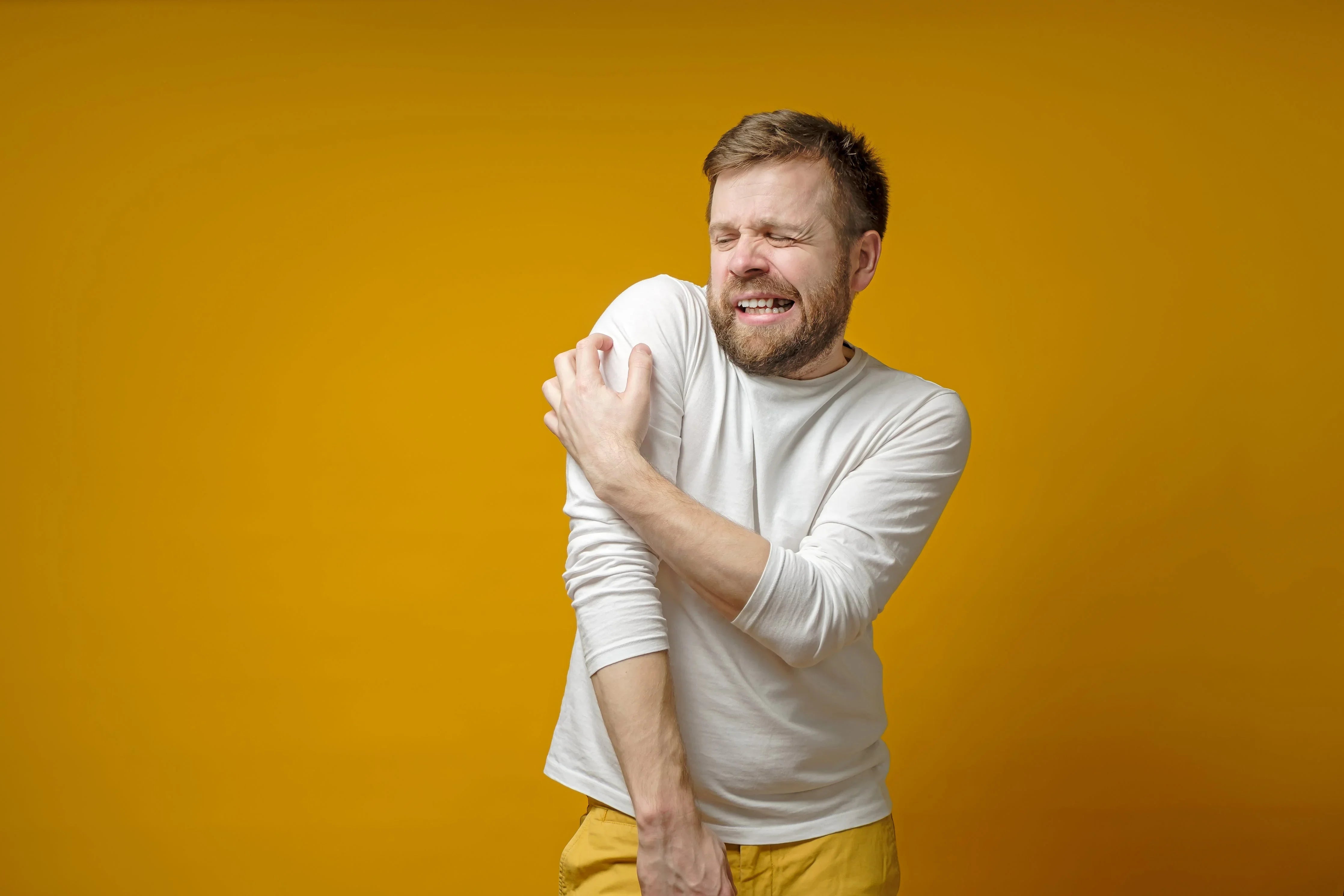 Man in white shirt scratching arm, possible allergy, on yellow background