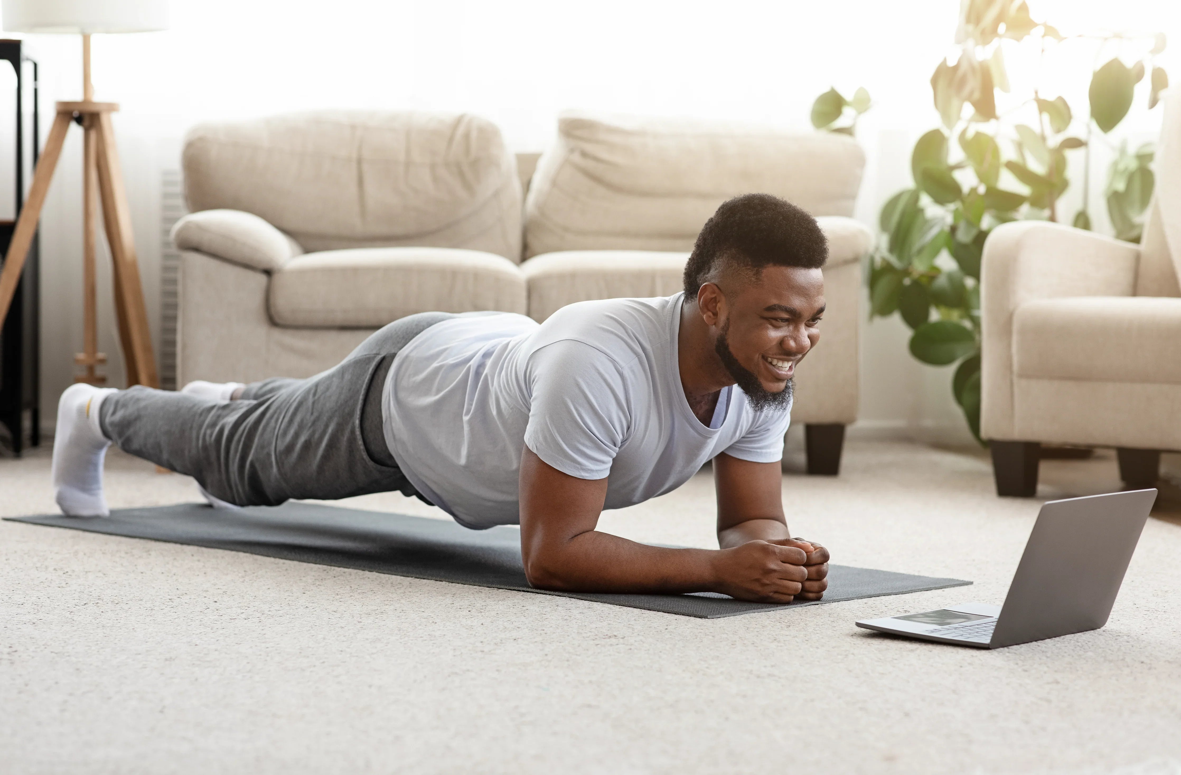Man in hypoallergenic cotton shirt and sweatpants doing plank exercise at home with laptop