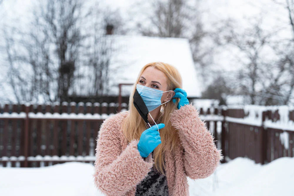 Woman in winter coat and gloves outdoors, adjusting face mask in snowy background