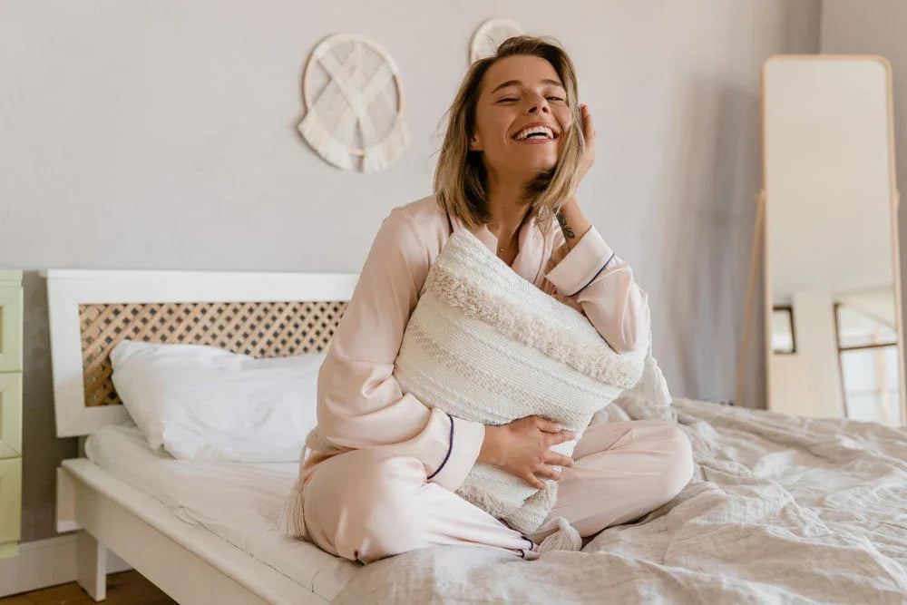 Smiling woman in pale hypoallergenic cotton pajamas sitting on a bed, hugging a pillow
