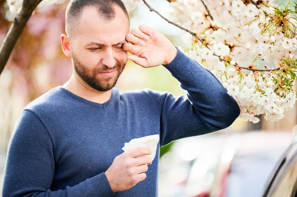 Man in blue allergy-free cotton sweater wiping eyes outdoors near blooming tree, holding tissue.
