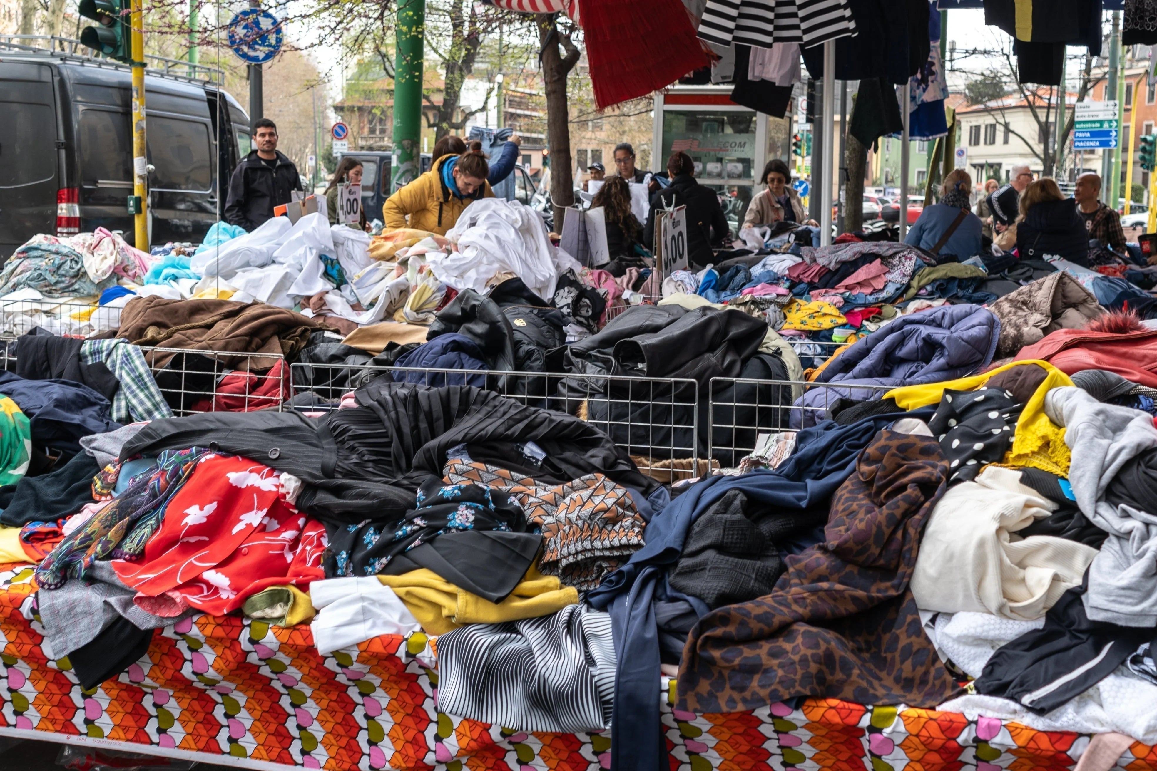 Outdoor market with piles of assorted clothing and people browsing for apparel deals