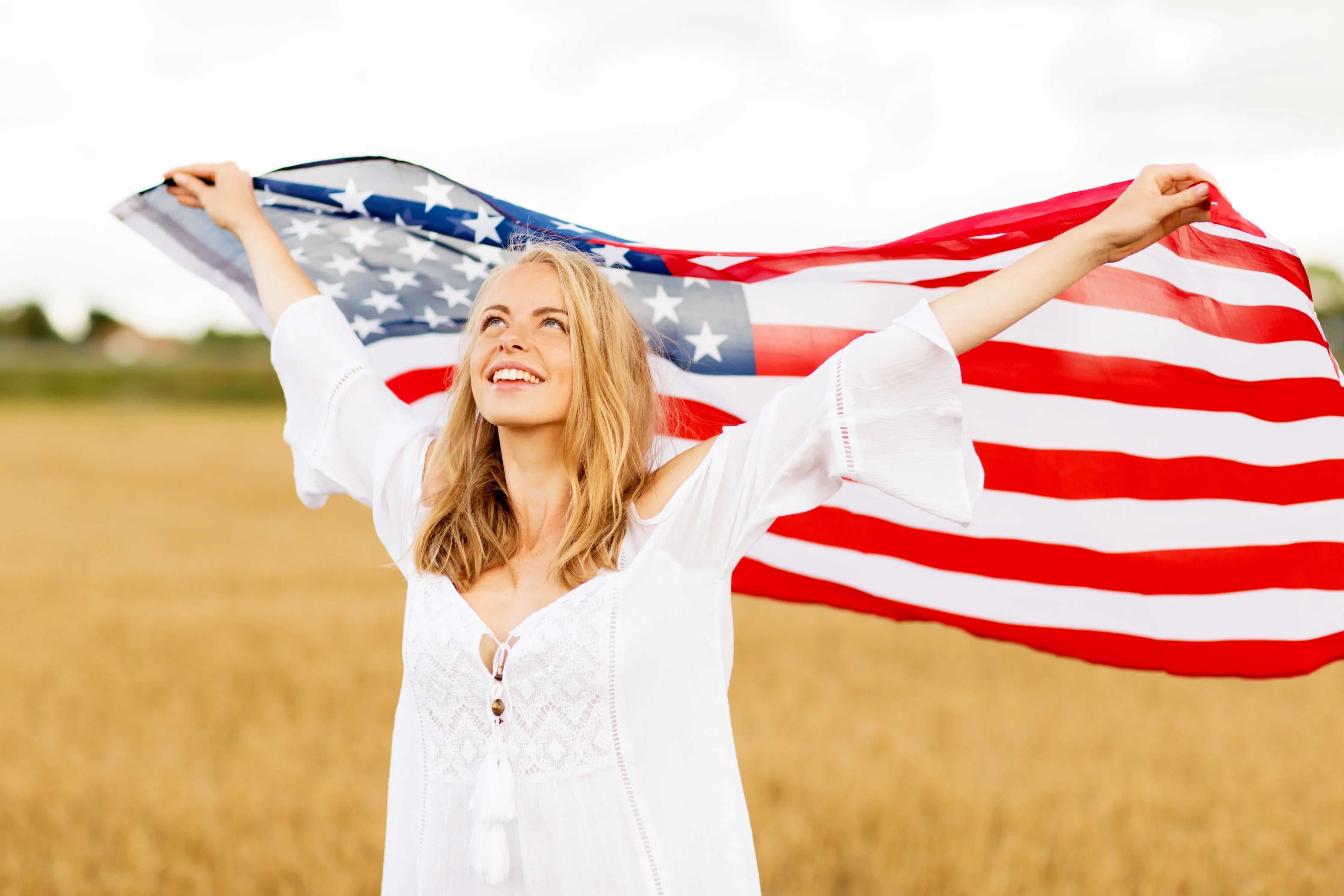 Woman in white dress holding American flag in field, smiling, natural cotton clothing