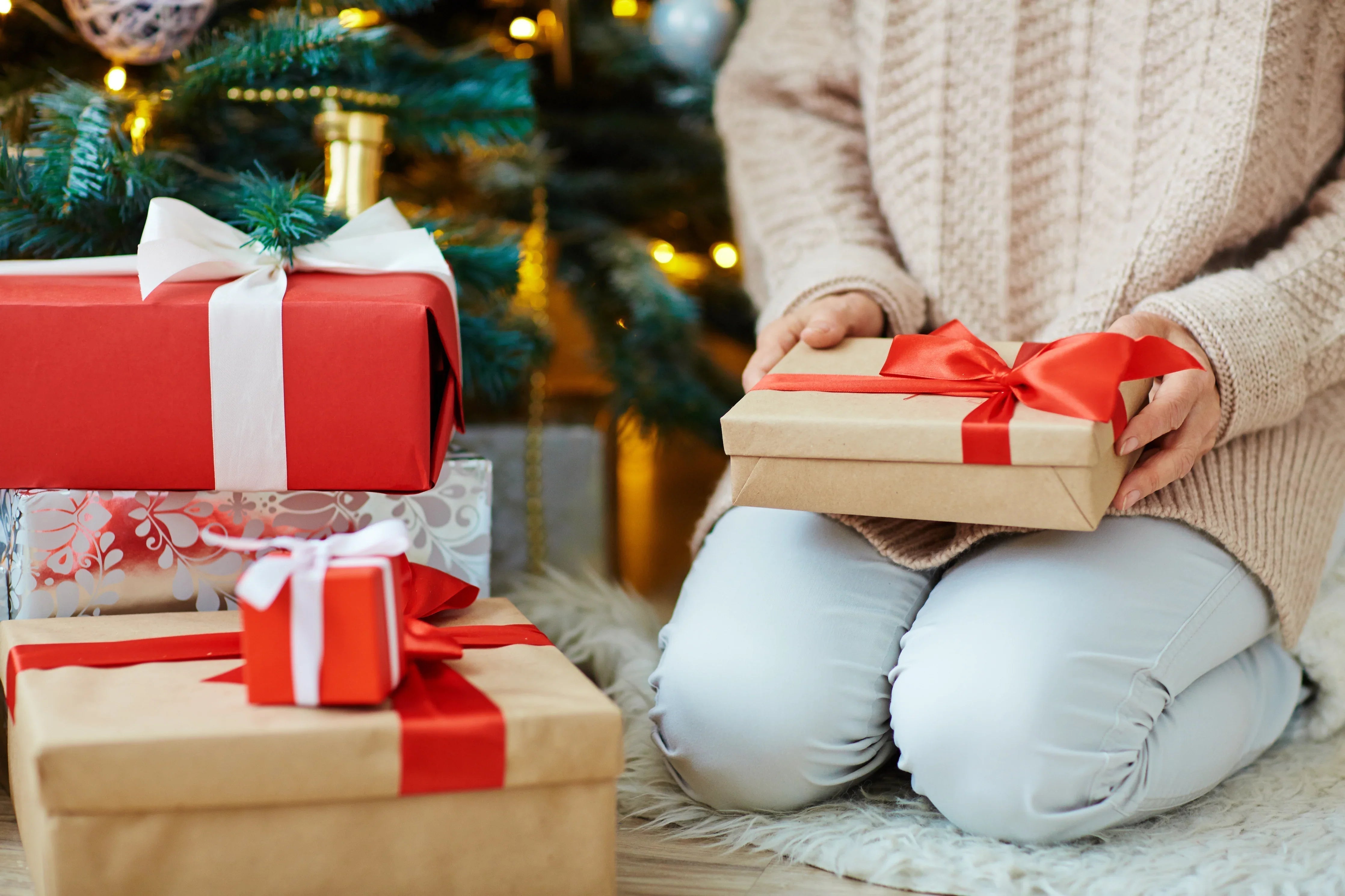 Person in knit sweater holding gift box with red ribbon beside Christmas tree and presents