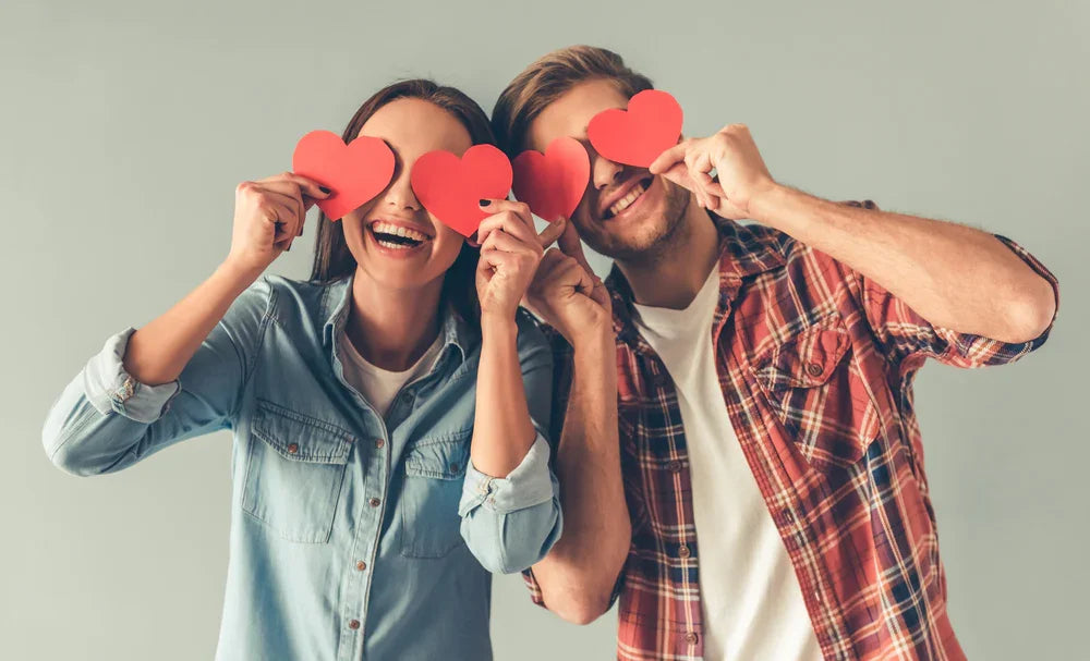 Smiling couple in casual, allergy-free cotton clothes holding red paper hearts to eyes