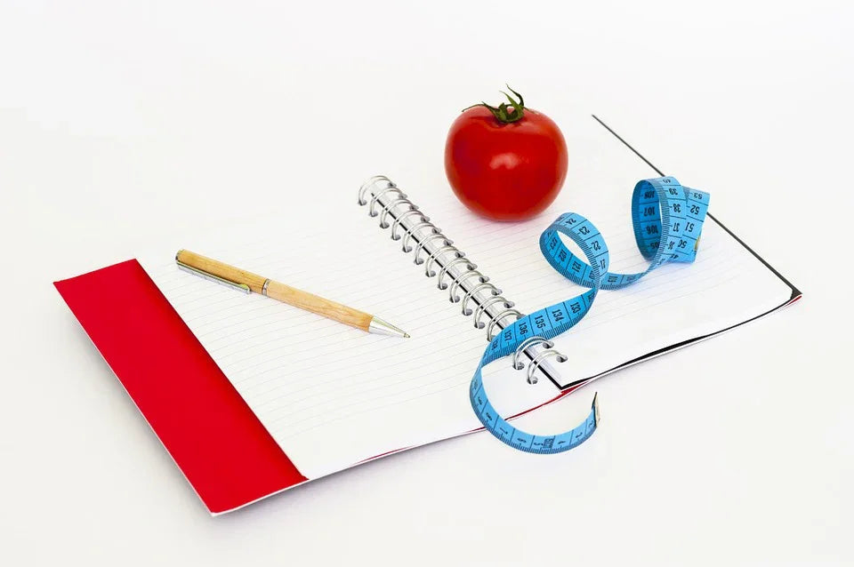 Open notebook with pen, blue measuring tape, and tomato on a white background, diet concept
