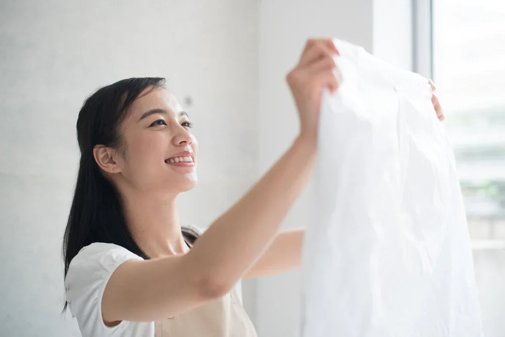 Smiling woman holding up a white organic cotton shirt near a bright window