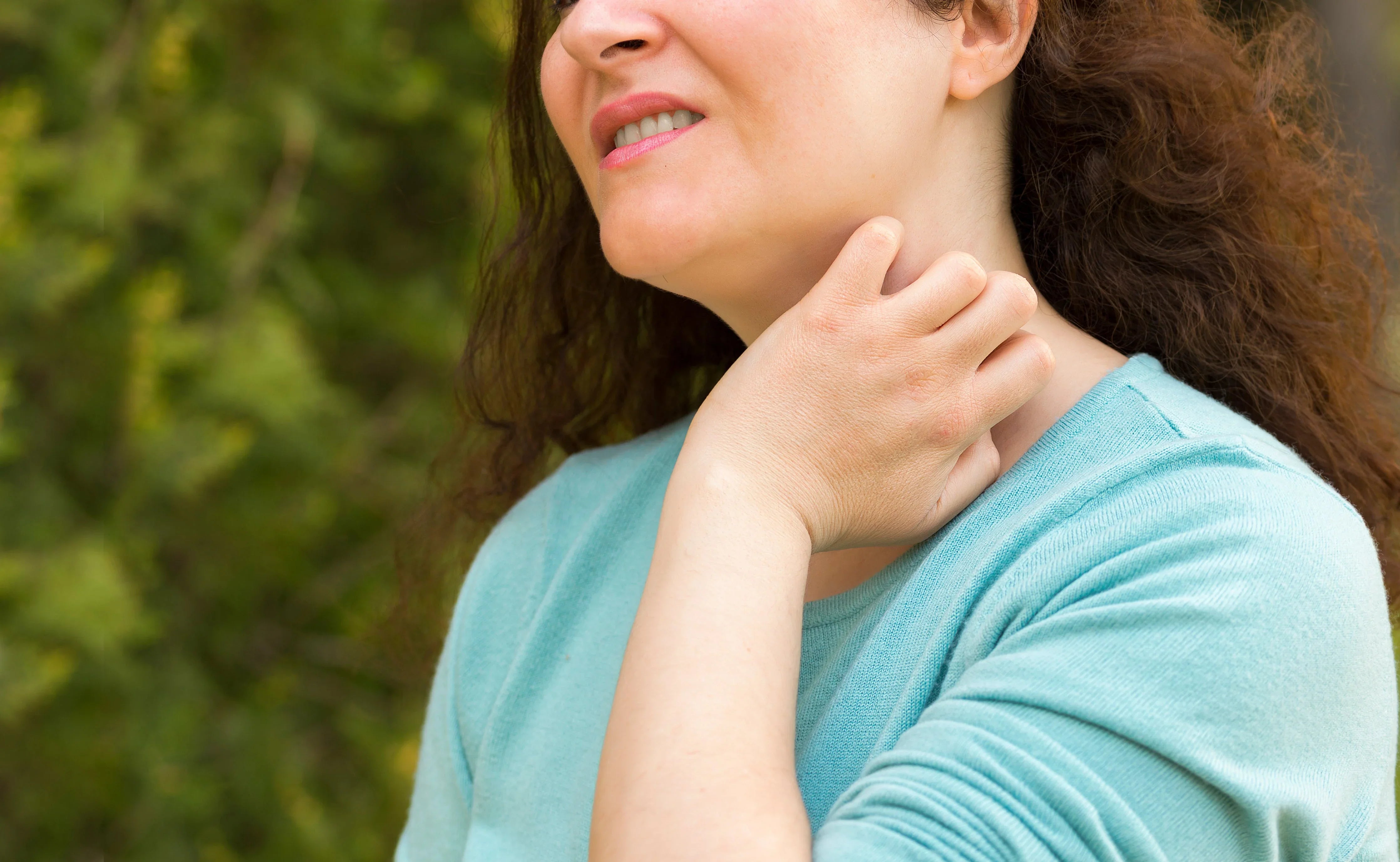 Woman in blue shirt scratching neck outdoors, showing skin irritation or allergy symptoms
