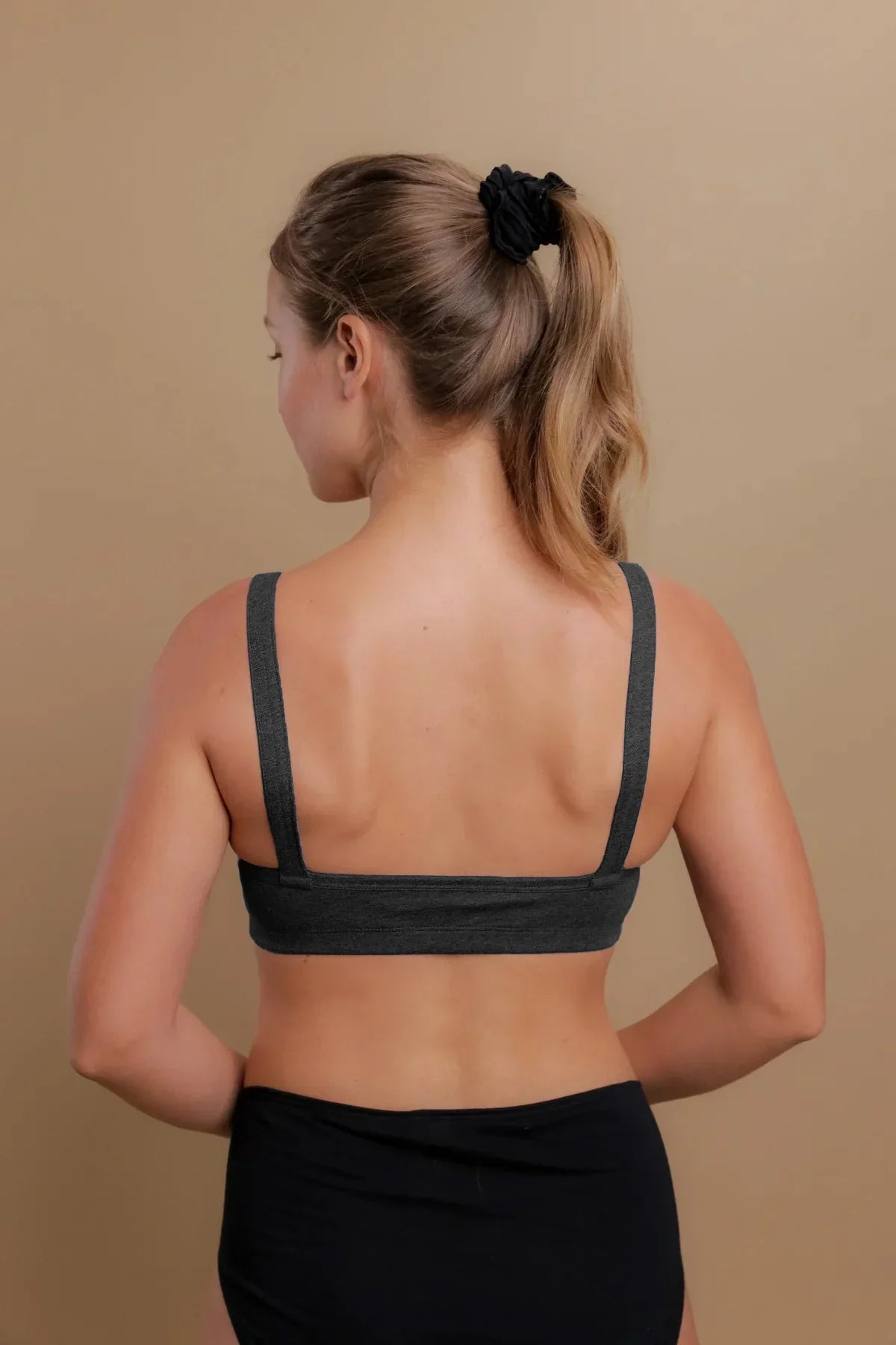Woman wearing dark grey hypoallergenic cotton bra, back view, against beige background